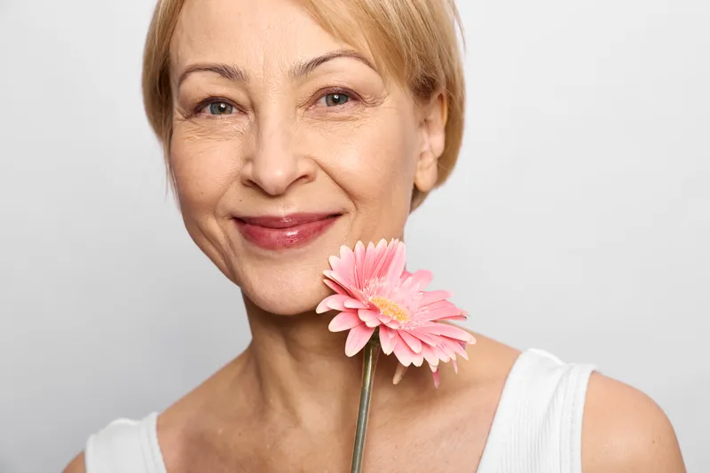 Woman portrait with gerbera smile, mature blonde female holds pink flower near lips, studio closeup on white background, calm happiness, fresh spring beauty