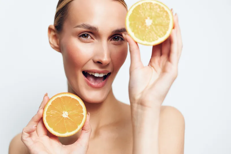 Cheerful woman with smooth skincare holding lemon slice near face, smiling in a clean studio portrait, fresh citrus aroma concept, bright natural light, minimal background