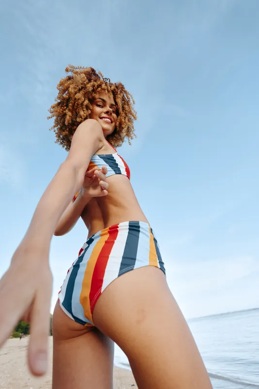 Smiling woman in bikini with curly hair posing on beach, ocean sea and blue sky behind, summer vacation mood, natural sunlight, relaxed posture, travel