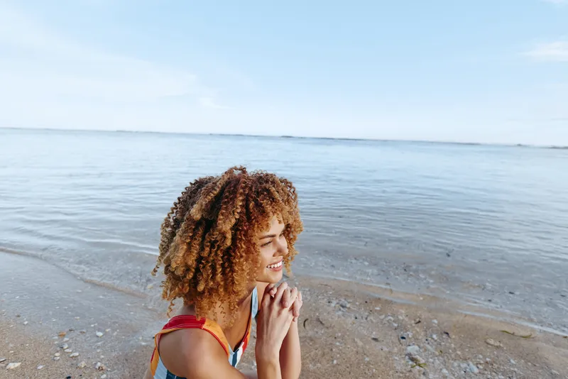Beach woman at ocean sea in summer relaxation curly hair smiling profile natural light on sandy shoreline soft waves calm outdoors wind blue sky travel mood wellness