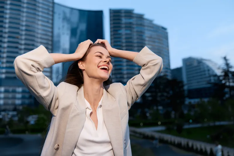 Energetic businesswoman celebrating success in an urban setting, wearing a stylish blazer and exuding joy with a modern cityscape backdrop.