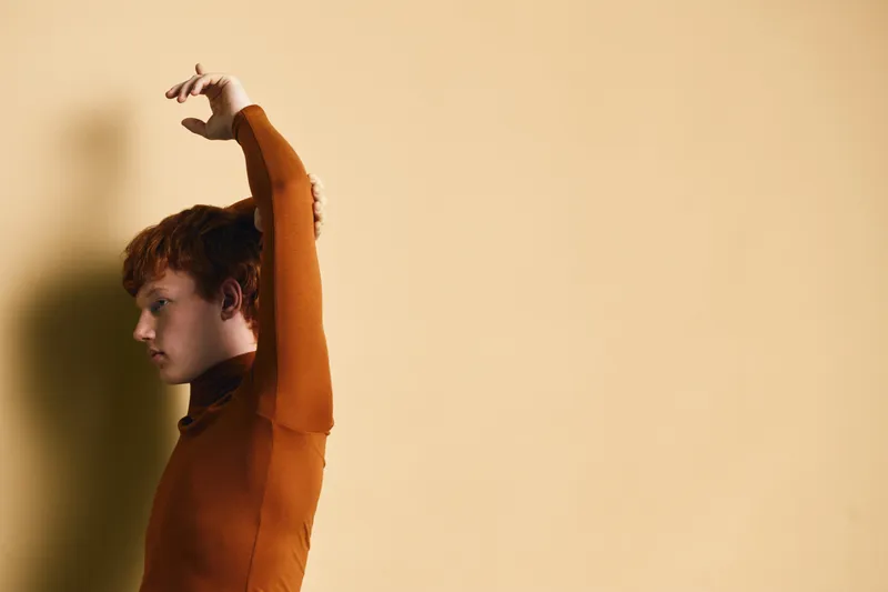 Young man with red hair stretching arms up against beige background in casual clothing. Side profile, motion and shadow on wall for relaxation and health.