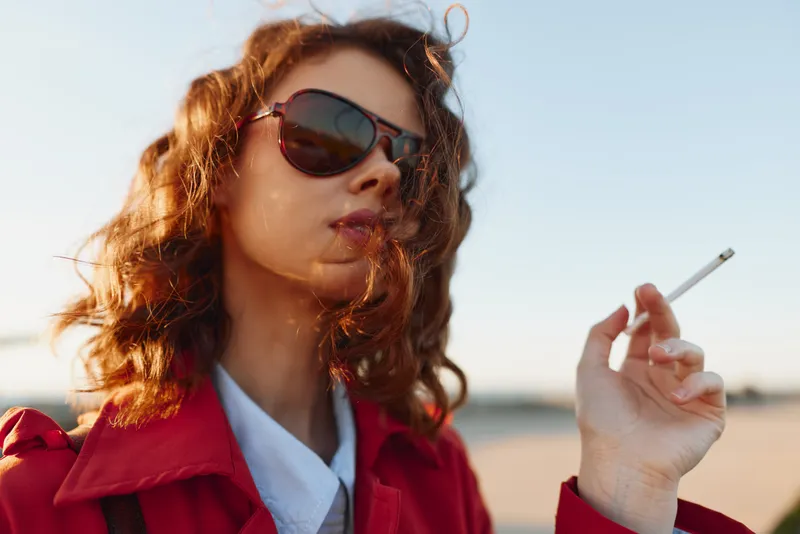 A stylish young woman with redhair and sunglasses stands on a sunlit beach, cigarette in hand as wind tousles her hair and the horizon glows softly.