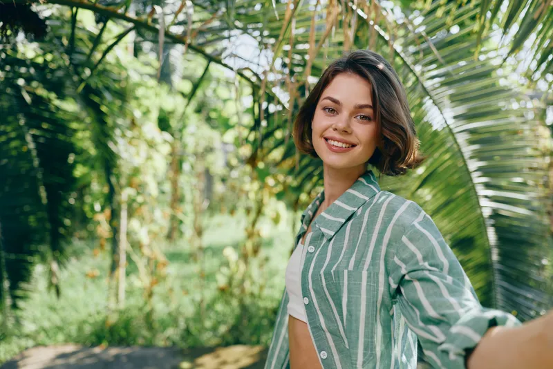 Portrait of a cheerful woman outdoors wearing a striped green jacket, smiling amid lush tropical plants.