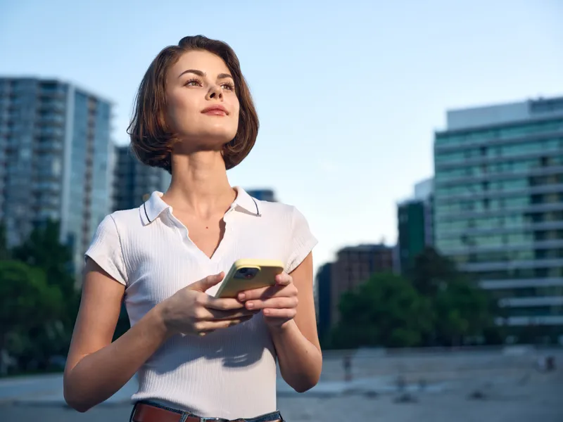 A poised young woman stands outdoors in a modern city environment, holding a smartphone. She gazes upward with confidence, conveying focus and a contemporary professional vibe.