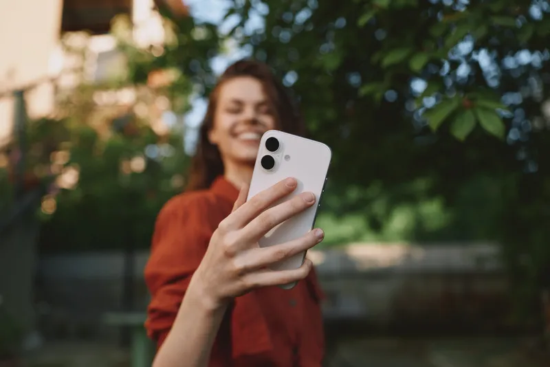 Happy woman takes a selfie outdoors with a smartphone, enjoying a casual lifestyle moment, warm sunlight and blurred greenery, relaxed pose, candid portrait for social media use