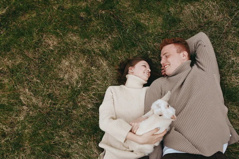 Young couple lies on grass smiling at each other while woman holds a small baby goat in cozy sweaters during a relaxed outdoor lifestyle moment.