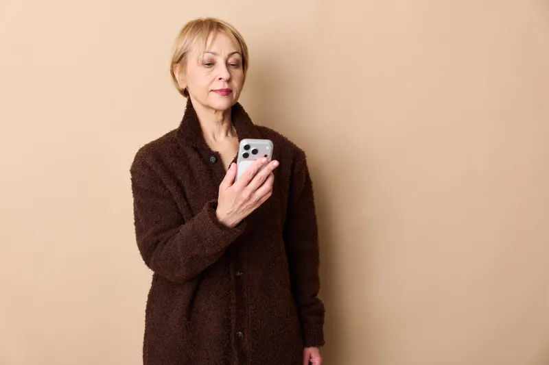 Elderly woman holds smartphone, looks worried and sad, shown alone against plain beige wall, loneliness and emotional stress, technology and communication concept for anxiety