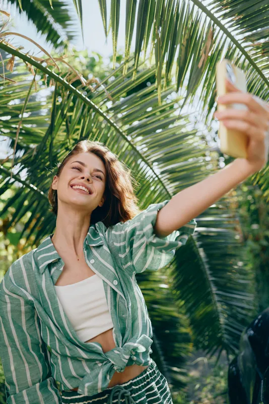 Cheerful woman taking a sunny selfie under lush palm leaves, wearing a green striped shirt and white top, capturing a candid, vibrant moment.