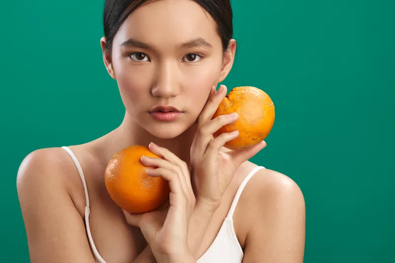 Asian woman holds fresh oranges in a bright studio setting, presenting a healthy, vibrant lifestyle with a calm, confident expression and colorful backdrop that conveys freshness and vitality.