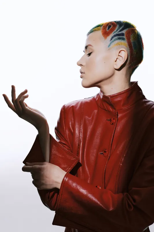 Woman with colorful painted hair in artistic patterns wearing red leather jacket posing with closed eyes and thoughtful expression on white background.