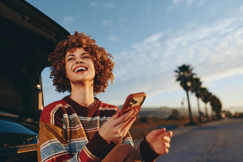 Woman lifestyle, smartphone, laughing, curly, hair, car, street, sunset, travel, outdoor portrait, seated by open car door, holding phone, smiling, urban palms, evening sky