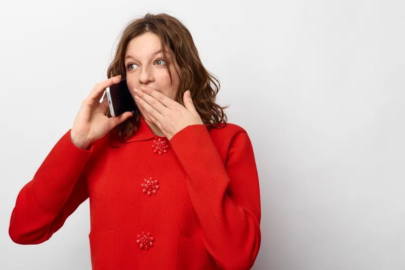 Young woman in red sweater talks on smartphone and covers mouth with hand, showing surprise. Isolated on white background. Concept of communication, emotion, and technology.