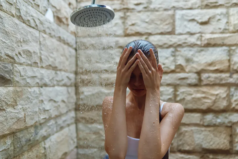 woman showering under outdoor rain shower with stone wall background, wet hair, relaxed mood, natural light, casual clothing, wellness concept