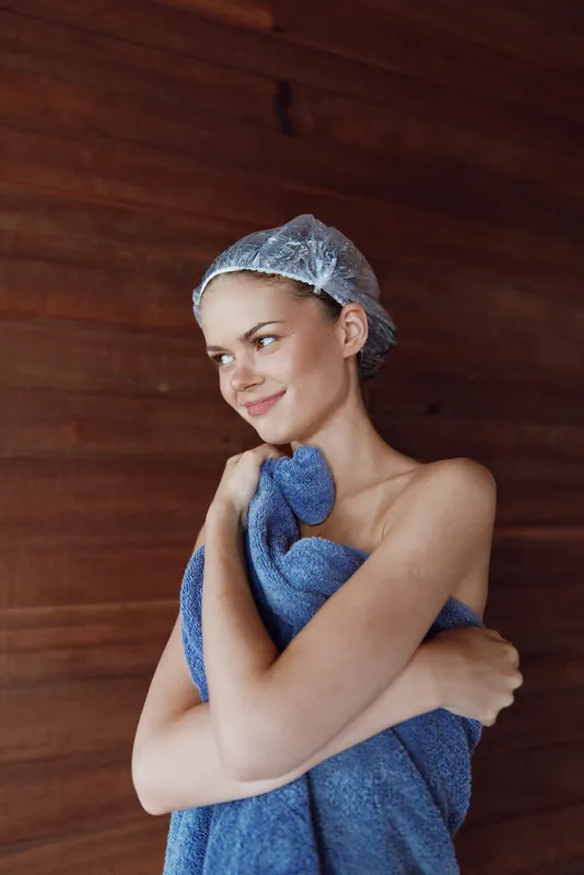 young woman wrapped in blue towel with shower cap smiling gently, wooden background, natural light, relaxed and fresh after bath or spa