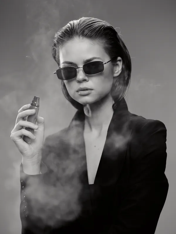 Black and white portrait of a woman with short hair wearing sunglasses and holding a lighter with smoke around her