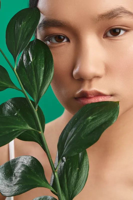 Asian woman gazes softly at the camera behind lush leaves in a clean studio setting, creating a calm, contemporary portrait that blends nature with refined beauty.