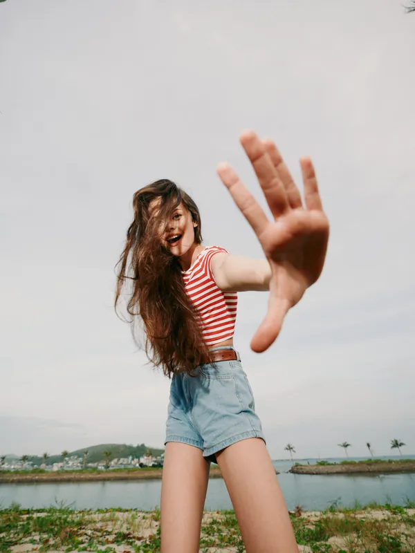 A playful woman in a striped crop top and denim shorts reaches toward the camera on a sunny beach with calm water and distant palm trees, exuding energy and spontaneity.