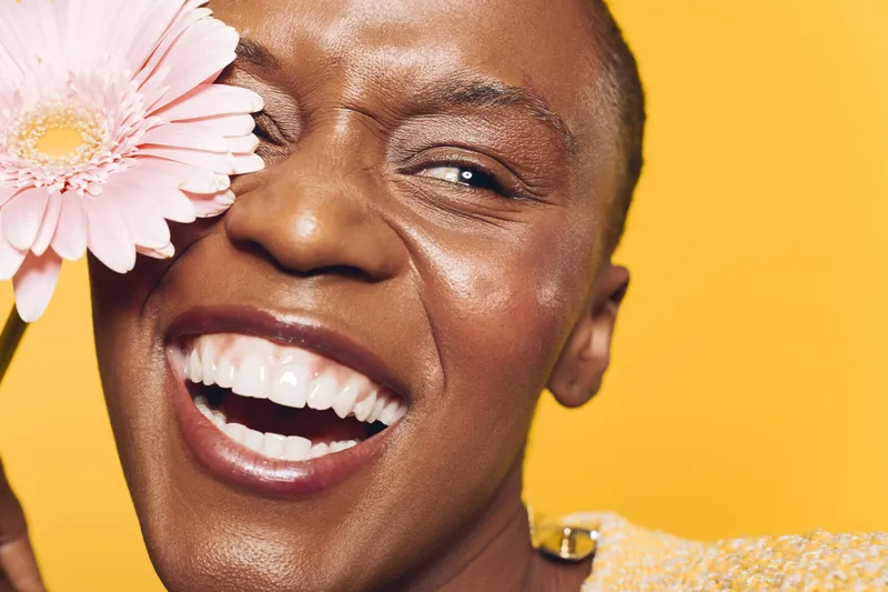 Happy African woman with dark skin smiles widely holding a pink flower near her face on yellow background. Concept of joy, beauty, natural skin, and positive emotions.