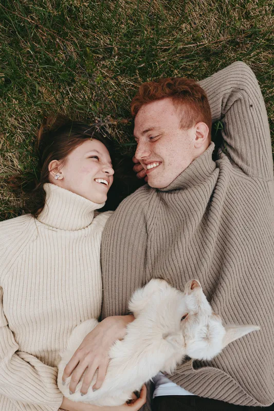 Young couple lying on grass in cozy sweaters smiling and holding a white baby goat together, enjoying a relaxed lifestyle moment outdoors in casual autumn clothing.