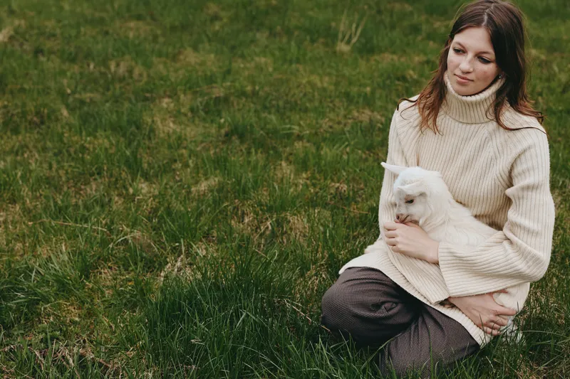 A young woman in a beige sweater sits on grass holding a small white baby goat, enjoying a peaceful moment outdoors in a natural lifestyle setting.