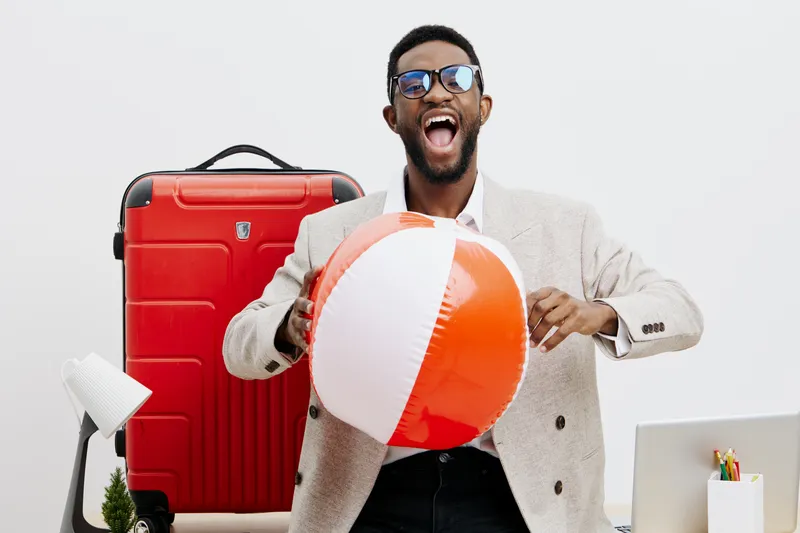 Cheerful young African-American man in sunglasses wearing a light blazer holding a large beach ball, with a red suitcase and white desk lamp in bright studio setting. Casual vacation travel concept.