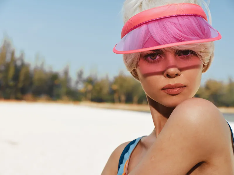 Portrait of a young woman at a sunny beach wearing a pink visor and swimsuit, vivid pink hair, clear skin, and soft sunlight highlighting facial features and a vibrant summer vibe.