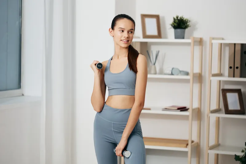 Woman in athletic wear holding a dumbbell during a workout in a bright home gym, focusing on fitness routine with natural light and calm atmosphere.