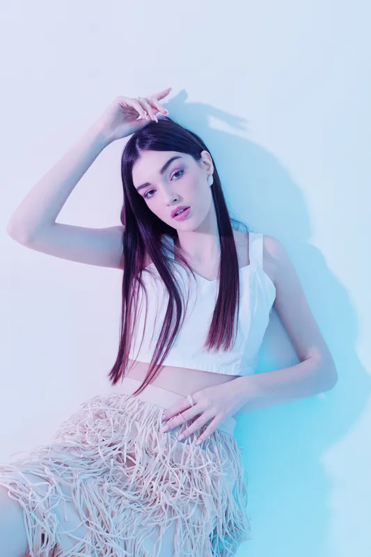 Young woman with long dark hair poses in a white top and textured skirt under soft blue and pink lighting. Concept of beauty fashion and modern style.
