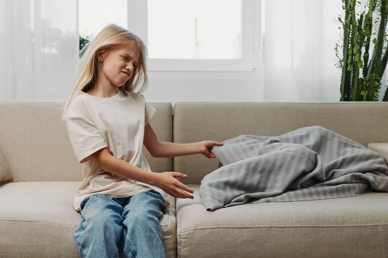 Little girl sitting on couch looks confused and frustrated at blanket with unusual shape in bright living room.