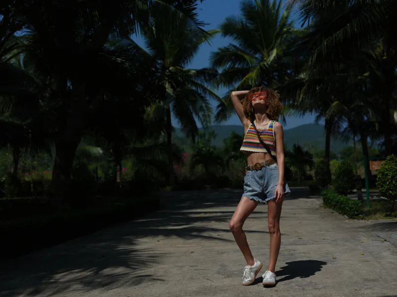 young woman wearing colorful striped top and denim shorts standing on sunny pathway surrounded by palm trees, enjoying warm weather and summer vibes