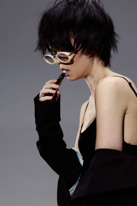 Stylish woman with short black hair wearing sunglasses and black outfit holds vape in thoughtful pose against gray background.