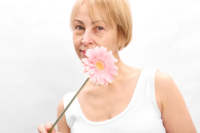 Elderly woman with pink flower closeup portrait, gentle smile, holding bloom on thin stem, soft studio light, white background, calm mood, minimal composition, sleeveless top