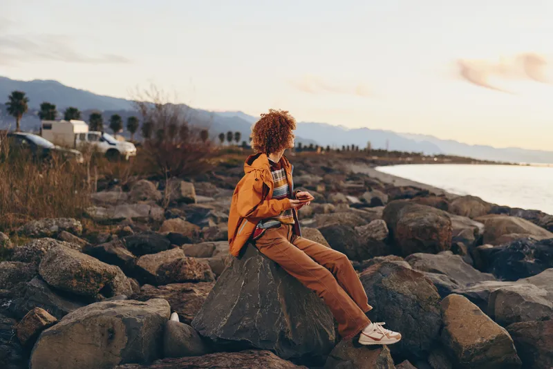 Woman lifestyle scene, mother sitting on rocks at beach sunset, holding child on her lap, relaxed travel moment, warm golden light, coastal landscape, quiet family bonding