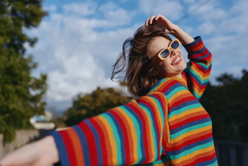 Woman in her mid twenties wearing a rainbow striped sweater and sunglasses, smiling outdoors in an urban park with arms out and wind in hair, joyful casual outfit for travel and lifestyle portrait.