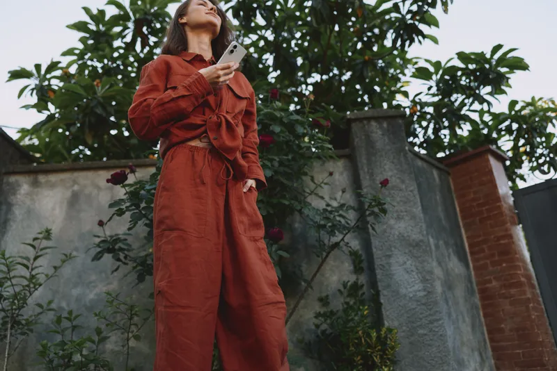 A woman in an orange jumpsuit stands outdoors, looking up while holding a smartphone. Casual street style portrait capturing contemporary fashion and everyday confidence