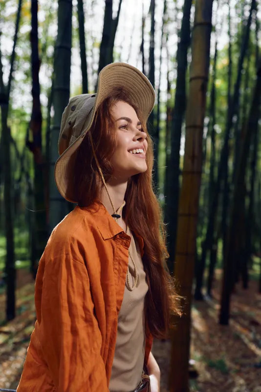 Woman smile hat bamboo forest outdoors portrait with long redhair and orange jacket, enjoying sunlit trail and peaceful nature, joyful profile during hiking and eco travel adventure.