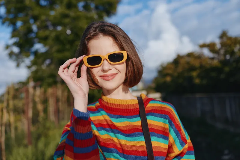 Woman, young woman, striped sweater, yellow sunglasses, smiling with short bob hair in a casual outfit outdoors; colorful knit, shoulder bag, sunlit urban park portrait for lifestyle travel.