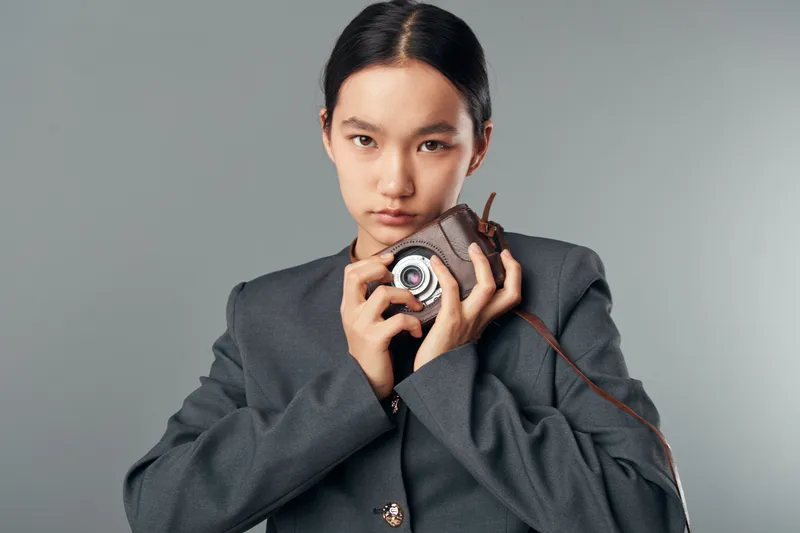 Young woman with serious expression holding vintage camera close to face for photography and creative concept on gray background in studio. Asian model in gray blazer.