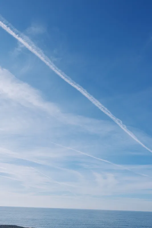 Blue sky with a long white contrail over calm ocean, distant horizon, minimal clouds, peaceful outdoor seascape under bright daylight.