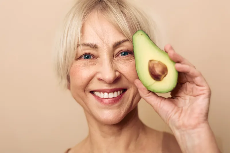 Smiling mature woman with short blonde hair holds half avocado near face. Natural skin care concept, healthy lifestyle, nutrition, wellness, and beauty. Studio portrait on beige background.