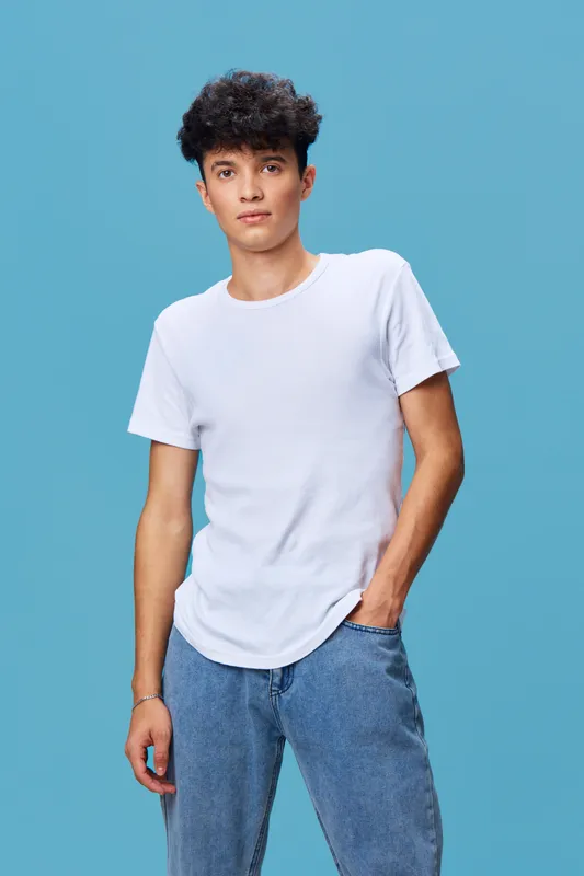 Young man in plain white t-shirt and jeans posing casually against a solid blue background with relaxed posture and neutral expression, showcasing casual fashion style.