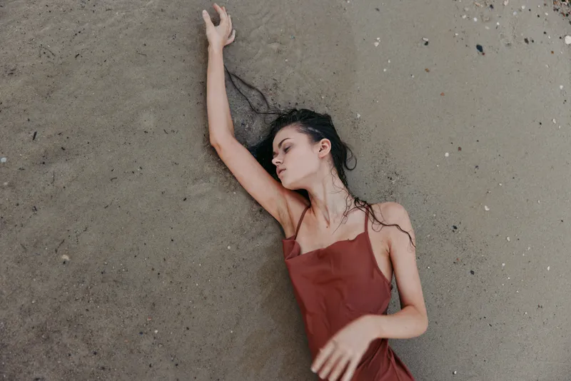 A solitary woman lies on the sandy shore in a burgundy dress, hair spread across the grain, a calm expression and soft light creating a minimal, intimate mood.