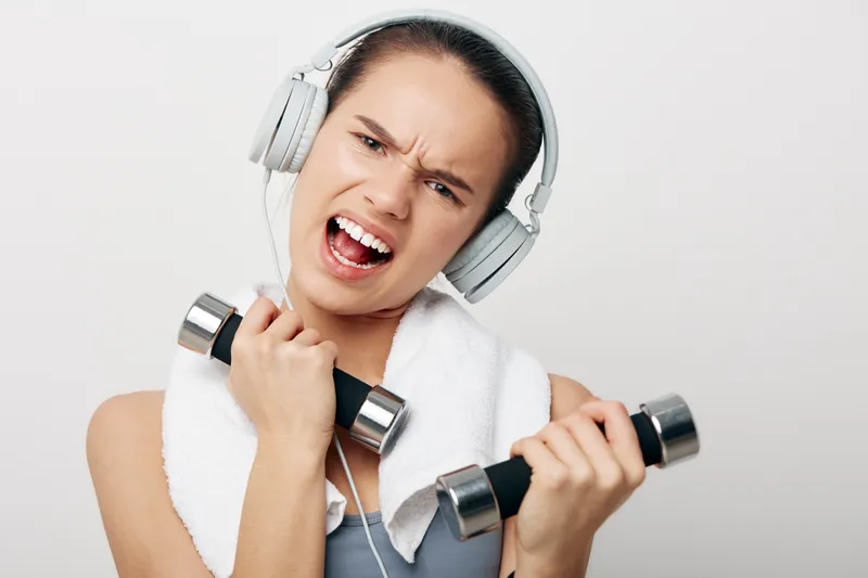 Woman in athletic gear lifts dumbbells with intense energy, wearing headphones and a towel around her neck, capturing a dynamic fitness workout moment.