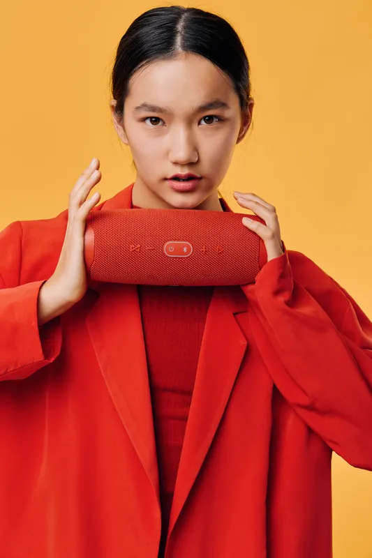 Woman in red coat holding portable wireless speaker with textured surface and buttons against yellow background. Stylish young female showing gadget with serious expression in studio.