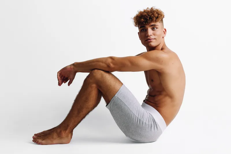 Young man with curly hair sitting on floor in gray shorts against white background, posing shirtless with relaxed expression and athletic body in studio setting.