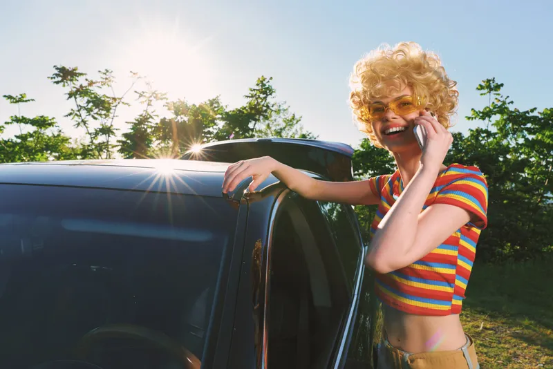 A woman in a striped crop top stands beside an open car window in bright sun, talking on a phone. Outdoor casual scene with vibrant colors conveys summer lifestyle, energy, and a cheerful road trip