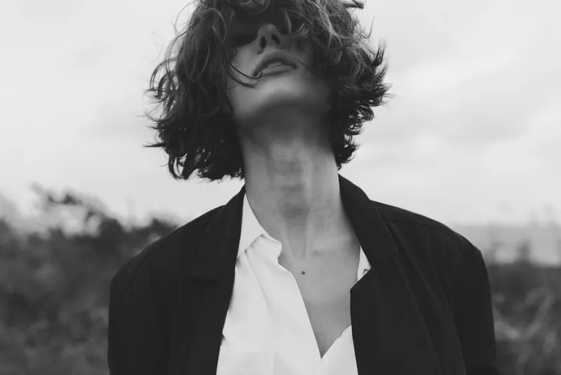 Moody black and white frame of a figure with tousled hair, open collar shirt and blazer, face partly hidden, atmospheric emotion, artistic mystery and introspective mood in a cinematic vibe