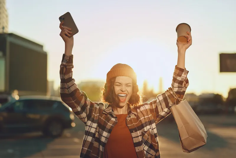 Woman celebrating outdoors with coffee, smartphone and shopping bag at golden hour, joyful candid portrait reflecting authenticity, mindful living and emotional storytelling in urban lifestyle.