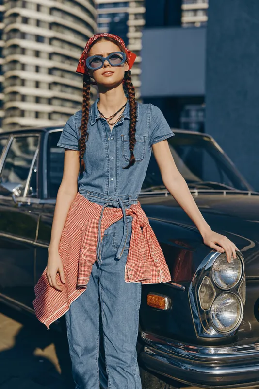 lifestyle woman in modern boho-western denim street style wearing sunglasses and a red checked shirt, film color tones with urban vintage car background